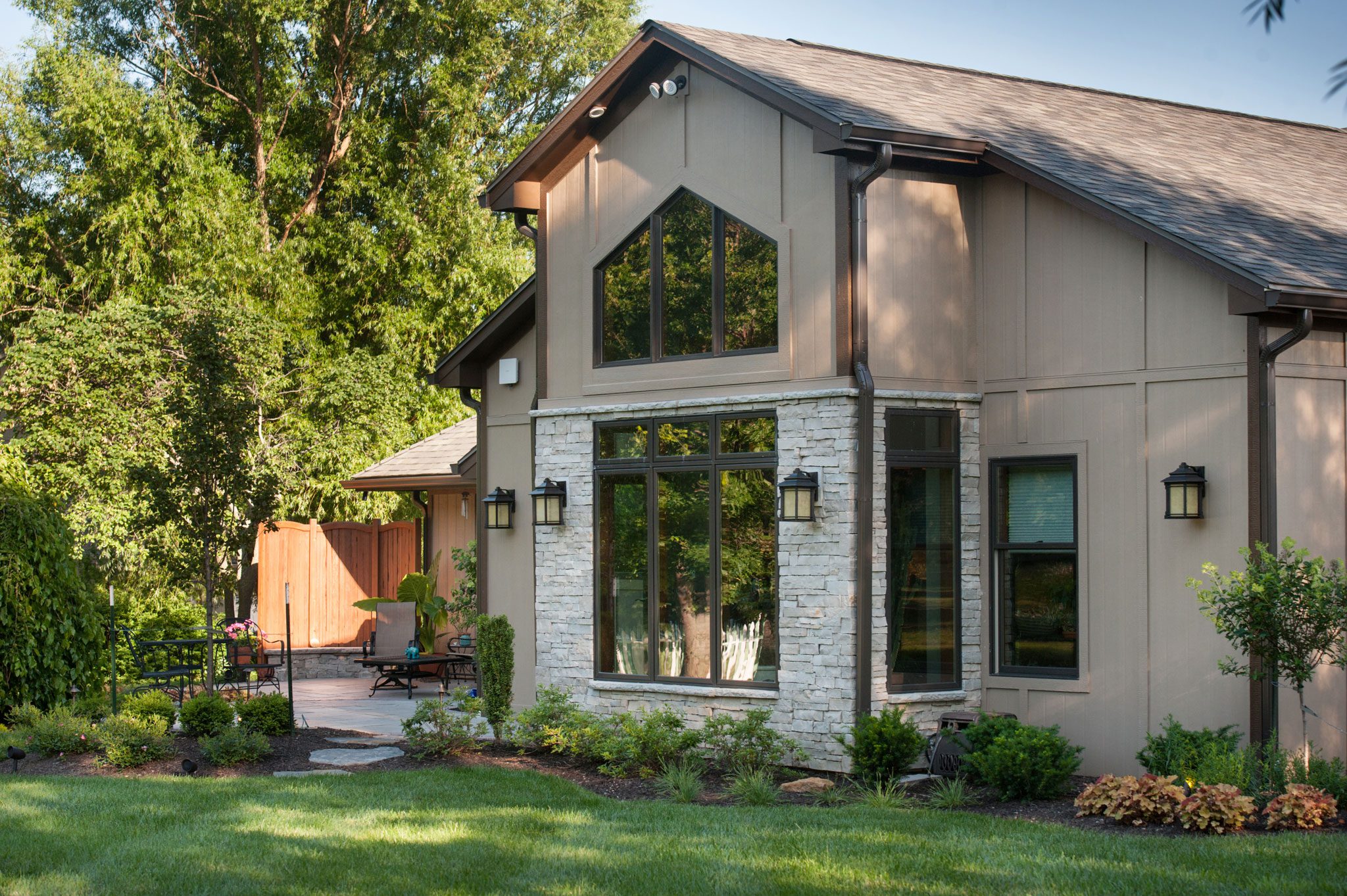 Modern house with large windows, stone accent wall, manicured lawn, lush garden, and patio furniture. Surrounded by trees and outdoor seating area.